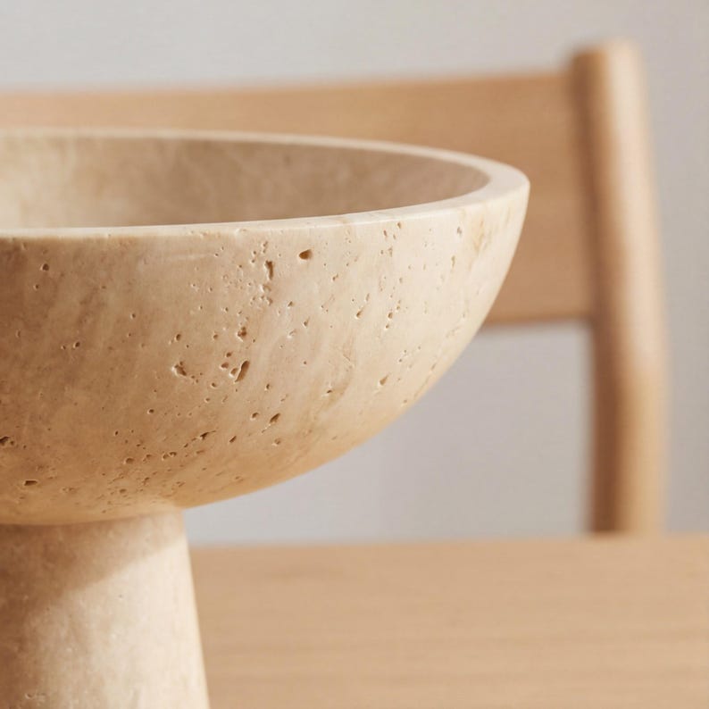 Puede incluir: Un cuenco beige texturizado sobre pedestal con una base ancha y poco profunda. El cuenco est&aacute; hecho de un material similar a la piedra y tiene forma redondeada. El fondo incluye una silla y una mesa de madera.
