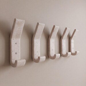 May include: A set of five off-white, curved wall hooks. Each hook is mounted with two screws and has a rounded, downward-facing hook. The hooks are made of a textured material and are mounted on a neutral-colored wall.