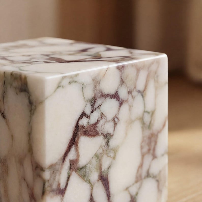 May include: A square marble block with a polished finish. The marble has a creamy white base with intricate burgundy and green veining, creating a natural pattern. The block's edges are clean and sharp, showcasing the stone's detail.