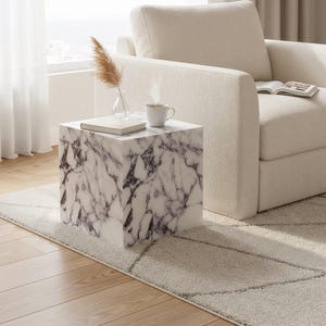 May include: A square marble-patterned side table with a book, a small vase with dried grass, and a white mug with steam rising from it. The table is next to a cream-colored armchair on a light-colored rug.