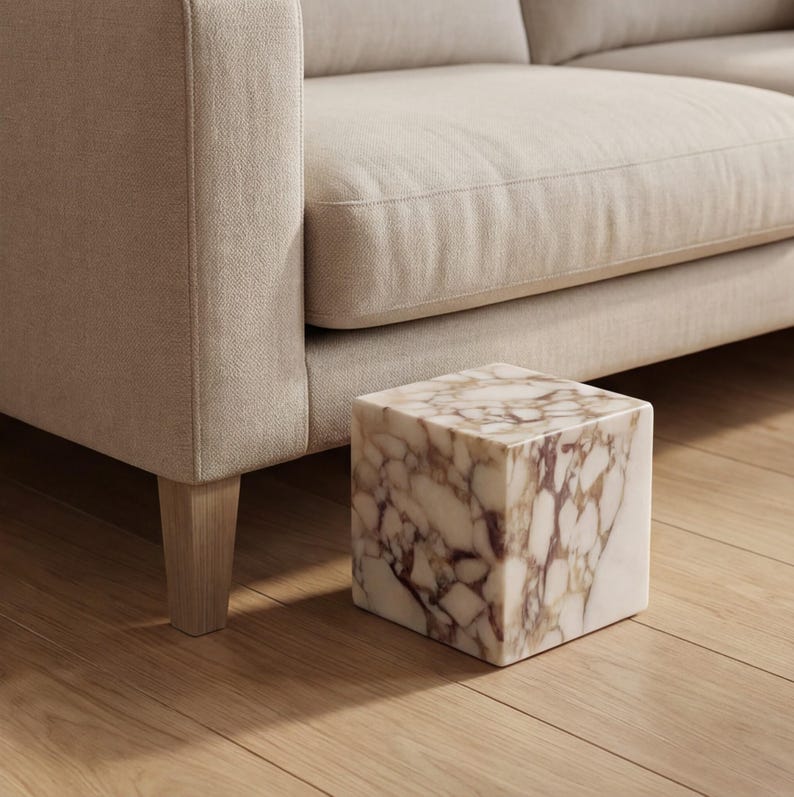 May include: A square marble side table with a cream and brown pattern sits on a wooden floor next to a beige sofa. The table is a cube shape, and the sofa has light-coloured upholstery.