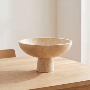 Puede incluir: Un cuenco de pedestal de piedra beige con una base ancha y poco profunda. El cuenco se asienta sobre una base cil&iacute;ndrica corta. El cuenco est&aacute; sobre una mesa de madera de color claro, con un fondo neutro.