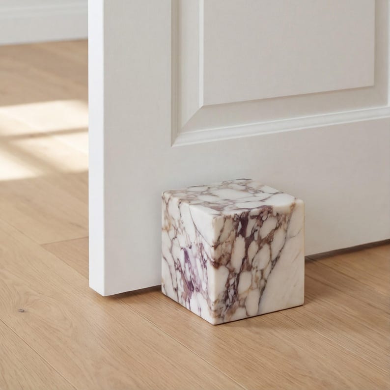 May include: A marble doorstop, cube-shaped, with a white base and purple veining. It's on a wooden floor next to a white door. The doorstop is approximately 10 cm square.