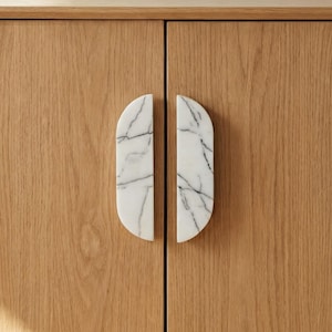 Puede incluir: Tiradores de puerta de armario ovalados de mármol blanco con vetas grises. Montados en un armario de madera clara. Estos tiradores son un elemento decorativo de ferretería.