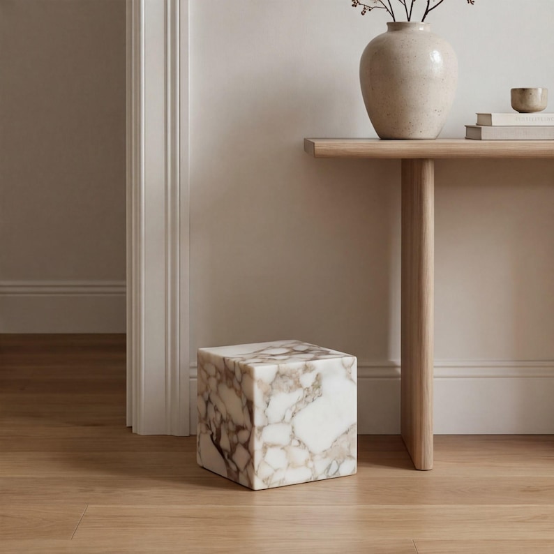 May include: A marble cube with white and brown marbling stands on a wooden floor. The square accent piece is near a light-coloured wooden table with a vase and books. The room has a neutral colour palette.