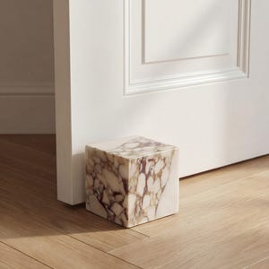 May include: A cube-shaped doorstop made of marble, displaying a pattern of white and brown veining. It is placed on a wooden floor, next to a white door. This decorative item is designed to hold a door open and is a home decor accessory.