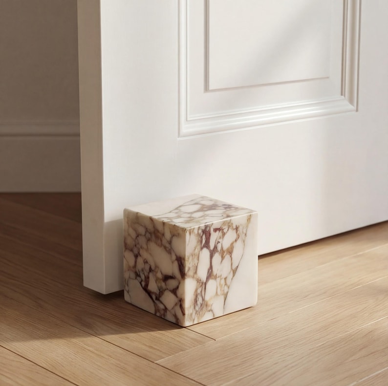 May include: A cube-shaped doorstop made of marble, displaying a pattern of white and brown veining. It is placed on a wooden floor, next to a white door. This decorative item is designed to hold a door open and is a home decor accessory.