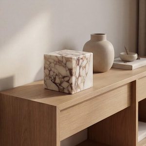 May include: A square marble decorative object with a brown and white pattern sits on a wooden surface. A beige vase and small bowl are also on the surface. The items are arranged in a minimalist style.