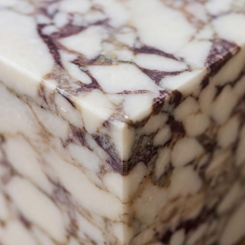 May include: Close-up of a polished marble block with a glossy finish. The marble features a white base with intricate patterns of deep purple and brown veining, creating a natural, elegant aesthetic. The edges are sharp and well-defined.