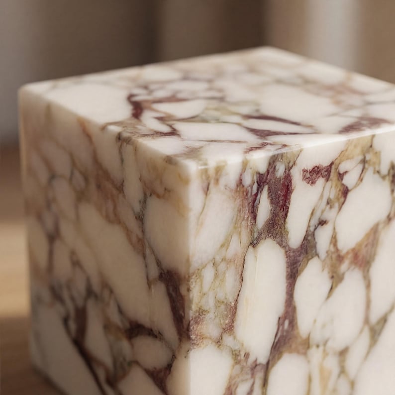 May include: A square marble block with a polished finish. The stone has a creamy white base with burgundy and tan veining, creating a natural pattern. The cube shape suggests a decorative or functional item, possibly a small table or plinth.