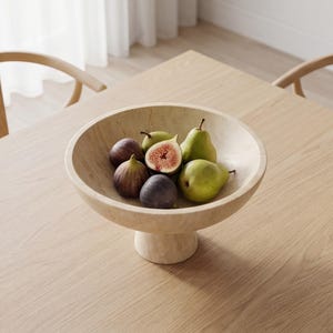 Puede incluir: Un cuenco de fruta beige con pedestal lleno de higos y peras frescas. El cuenco es de piedra de color claro y se asienta sobre una base corta y redonda. Los higos son morados y las peras verdes.