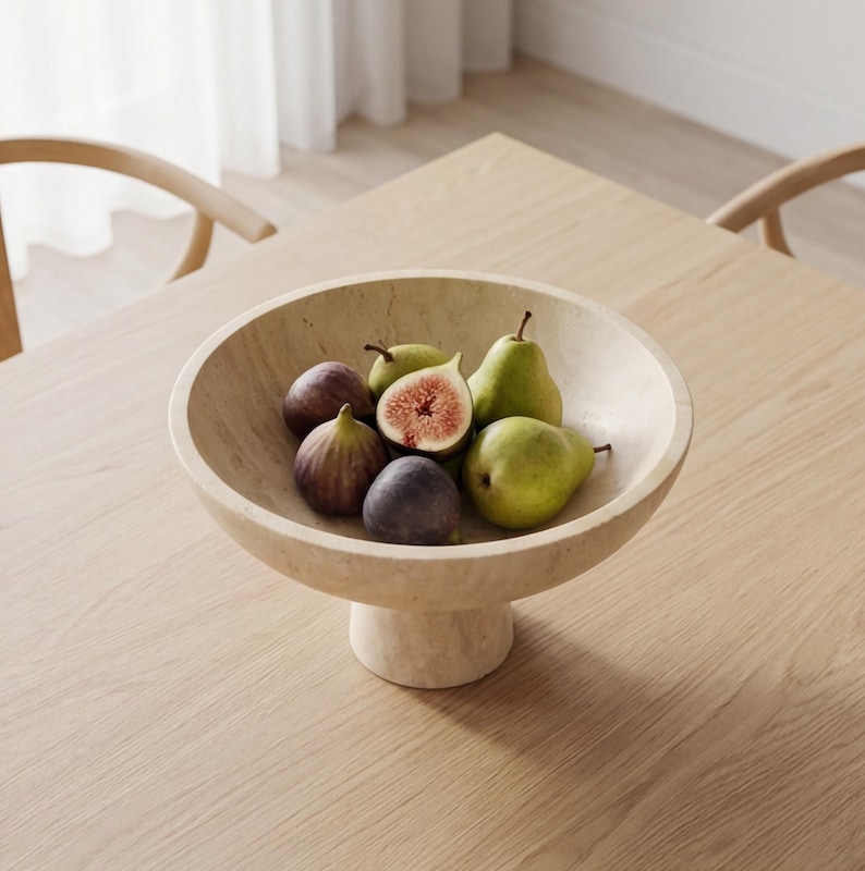 Puede incluir: Un cuenco de fruta beige con pedestal lleno de higos y peras frescas. El cuenco es de piedra de color claro y se asienta sobre una base corta y redonda. Los higos son morados y las peras verdes.