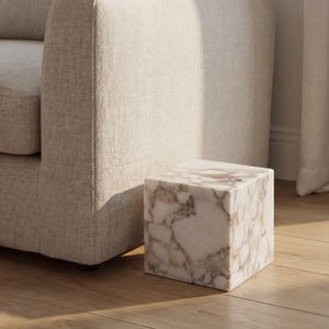 May include: A square marble side table with a white and tan veined pattern. The cube-shaped table is positioned next to a beige upholstered sofa on a hardwood floor. The table is approximately 45 cm wide.