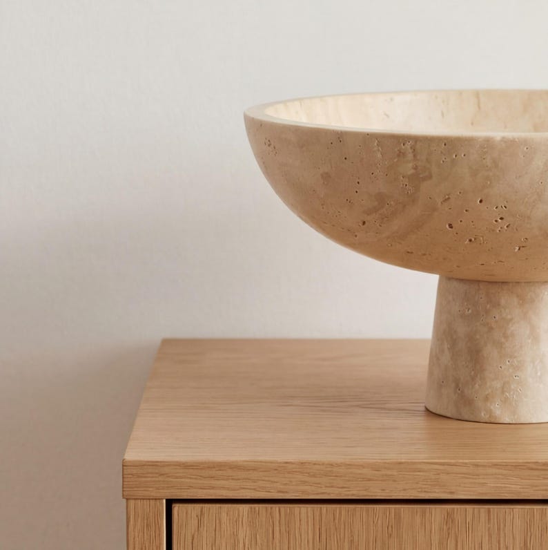 Puede incluir: Un cuenco de pedestal de piedra beige con una base ancha y poco profunda. El cuenco se asienta sobre una base corta y cil&iacute;ndrica. El cuenco y la base tienen una superficie texturizada y se exhiben sobre una superficie de madera de color claro.