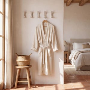 May include: A cream-colored robe hangs on a wooden hanger against a light-colored wall. A woven basket sits on a wooden stool. The room has a neutral color palette with a bed in the background.