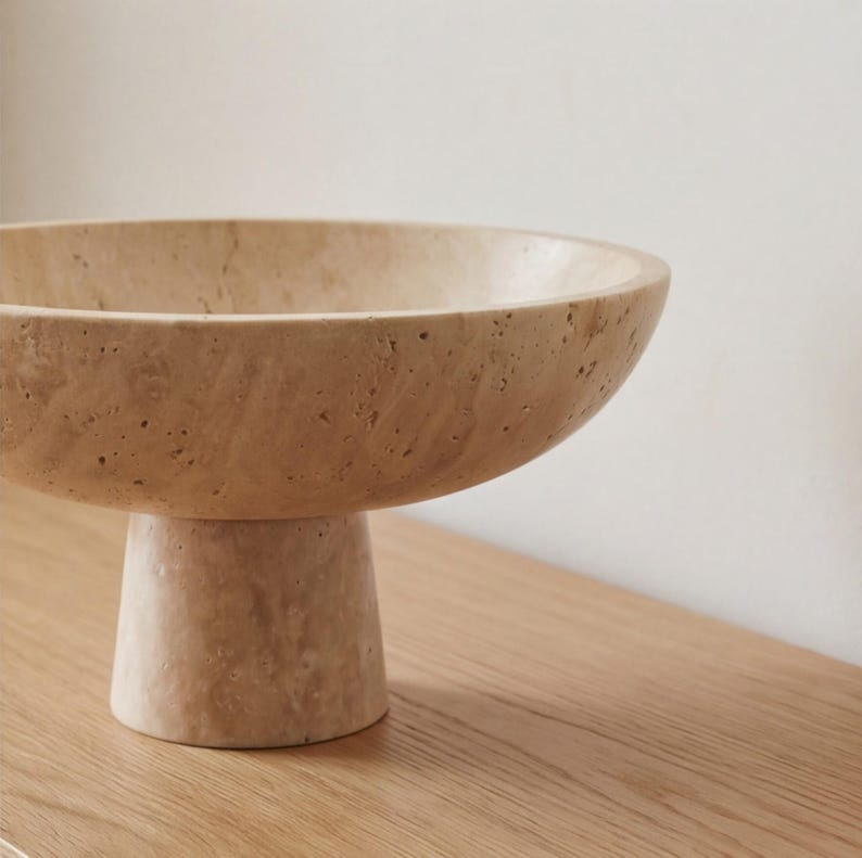 Puede incluir: Un cuenco de pedestal de piedra beige con una base ancha y poco profunda. El cuenco se asienta sobre una base cil&iacute;ndrica corta. La superficie tiene un aspecto texturizado y poroso, y el cuenco se exhibe sobre una superficie de madera.