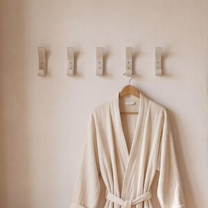 May include: A cream-colored robe hangs on a wooden hanger below five light-colored wall hooks. The robe is made of a soft, textured fabric and has a matching belt. The hooks are evenly spaced on a light-colored wall, creating a minimalist aesthetic.
