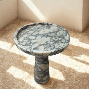 May include: A round marble side table with a pedestal base. The tabletop has a raised edge and a speckled pattern of white, grey, and black. The base is a solid, cylindrical shape with a matching marble pattern. The table is approximately 61 cm in height.