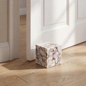 May include: A cube-shaped doorstop made of marble, featuring a white and purple veined design. It rests on a light wooden floor, beside a white door and frame. This decorative item is a functional home accessory, designed to hold a door open.