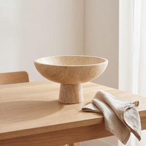 Puede incluir: Un cuenco claro sobre pedestal de piedra sobre una mesa de madera. Una servilleta de lino plegada de color claro descansa junto al cuenco. El cuenco tiene un dise&ntilde;o ancho y poco profundo.