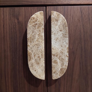 Puede incluir: Dos tiradores de armario semicirculares hechos de mármol beige pulido con vetas blancas intrincadas. Los tiradores están montados verticalmente en una puerta de armario de madera marrón oscuro, colocados lado a lado para formar un círculo.