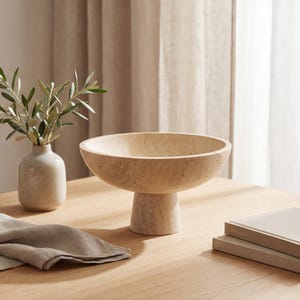 Puede incluir: Un cuenco beige sobre pedestal con una base ancha y poco profunda. El cuenco est&aacute; hecho de un material texturizado y se asienta sobre una base cil&iacute;ndrica corta. Un peque&ntilde;o jarr&oacute;n con ramas de olivo y una servilleta doblada tambi&eacute;n est&aacute;n sobre la mesa de madera.
