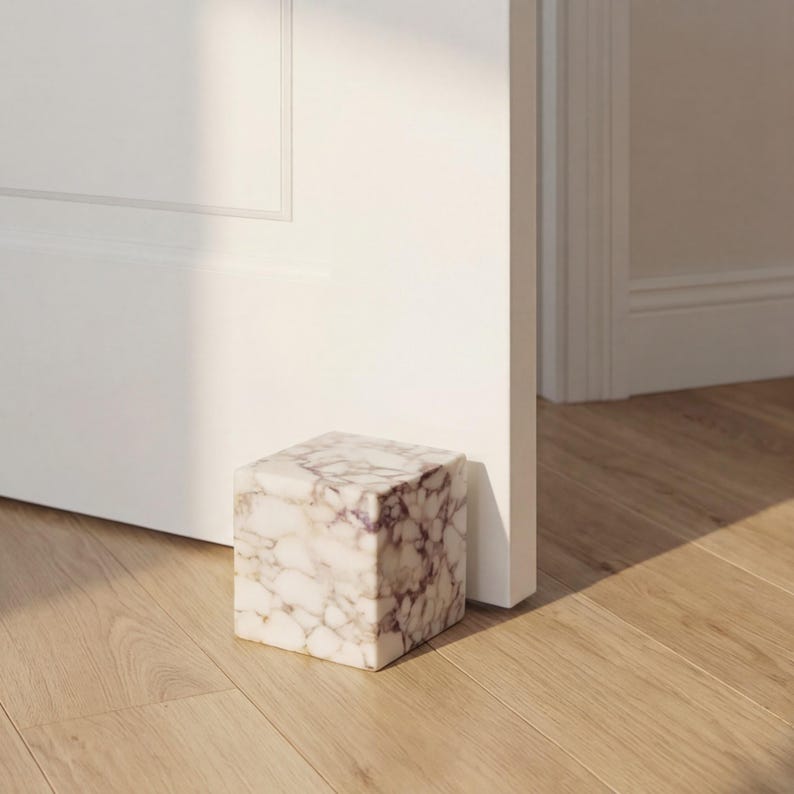 May include: A square marble doorstop with a white and purple veined pattern. The doorstop is on a wooden floor, holding a white door open. The doorstop is a decorative and functional home decor item.