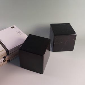 Puede incluir: Dos cubos de mármol negro con sutiles vetas blancas. Los cubos están sobre una superficie blanca junto a una pila de libros con cubiertas blancas. Los cubos tienen una superficie pulida y reflectante.