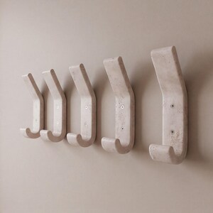 May include: Five off-white, wall-mounted coat hooks. Each hook has a curved design with a rounded bottom and a flat top. The hooks are mounted on a light beige wall. The hooks are made of a textured material.