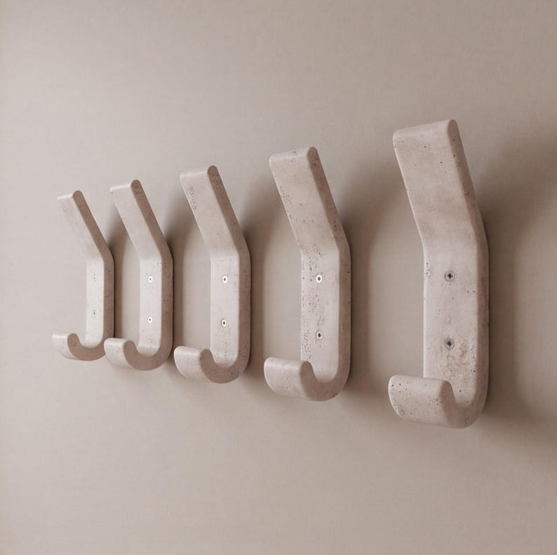 May include: Five off-white, wall-mounted coat hooks. Each hook has a curved design with a rounded bottom and a flat top. The hooks are mounted on a light beige wall. The hooks are made of a textured material.