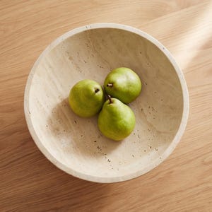 Puede incluir: Un cuenco redondo de piedra beige con una superficie texturizada contiene tres peras verdes. El cuenco est&aacute; sobre una superficie de madera. Las peras est&aacute;n maduras y listas para comer.
