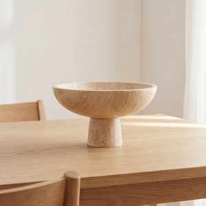 Puede incluir: Un cuenco beige con pedestal y una base ancha y poco profunda. El cuenco se asienta sobre una base cil&iacute;ndrica corta. El cuenco y la base tienen un aspecto texturizado similar a la piedra. El cuenco se exhibe sobre una mesa de madera clara.