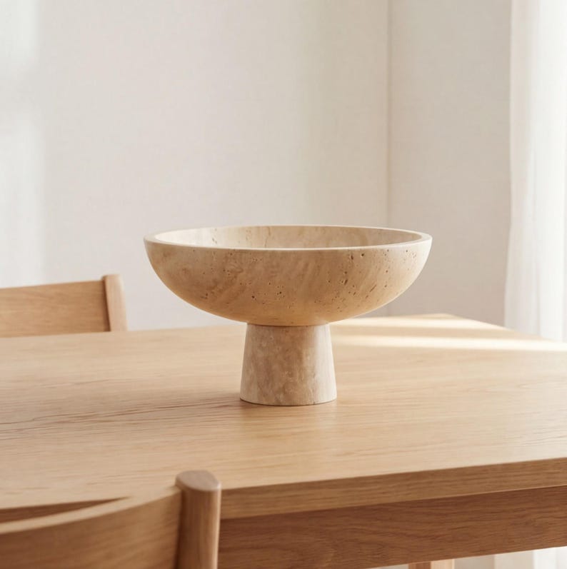 Puede incluir: Un cuenco beige con pedestal y una base ancha y poco profunda. El cuenco se asienta sobre una base cil&iacute;ndrica corta. El cuenco y la base tienen un aspecto texturizado similar a la piedra. El cuenco se exhibe sobre una mesa de madera clara.