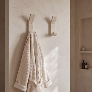 May include: Two beige stone wall hooks with a waffle-textured robe hanging on one. The hooks are mounted on a light beige wall. The robe is a light cream color and has a matching belt. The bathroom setting has a minimalist design.