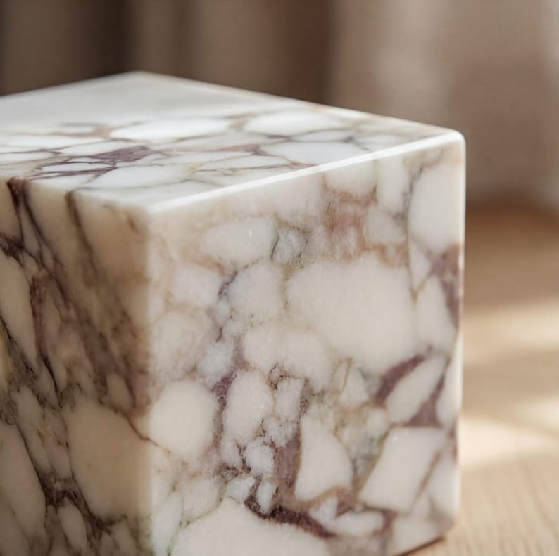 May include: A square marble block with a polished surface. The marble has a white base colour with veining in shades of burgundy and grey, creating a natural pattern. The block's edges are clean and sharp, showcasing the stone's smooth texture.