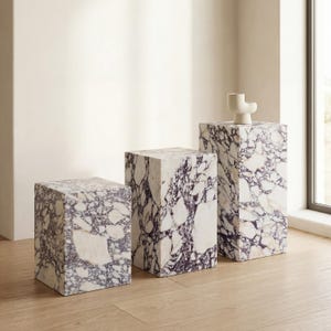 May include: Three marble pedestals of varying heights. The pedestals are cube-shaped and feature a white and purple veined pattern. A small, white, abstract sculpture sits atop the tallest pedestal. The pedestals are arranged on a light-colored wooden floor.