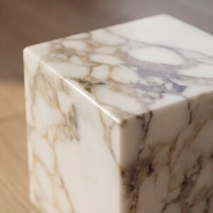May include: Close-up of a marble cube with a polished finish. The cube has a white base with brown and grey veining. The edges are smooth and the surface is reflective.