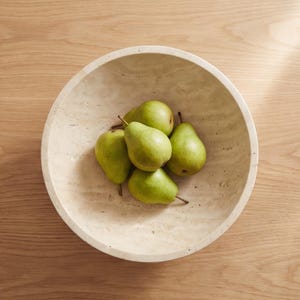 Puede incluir: Un cuenco de piedra beige lleno de seis peras verdes. El cuenco tiene forma redondeada y est&aacute; sobre una superficie de madera marr&oacute;n claro. Las peras est&aacute;n maduras y tienen tallos marrones.
