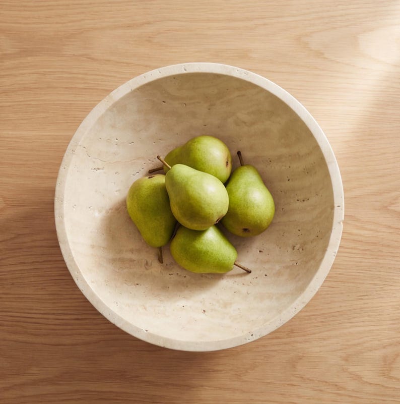 Puede incluir: Un cuenco de piedra beige lleno de seis peras verdes. El cuenco tiene forma redondeada y est&aacute; sobre una superficie de madera marr&oacute;n claro. Las peras est&aacute;n maduras y tienen tallos marrones.