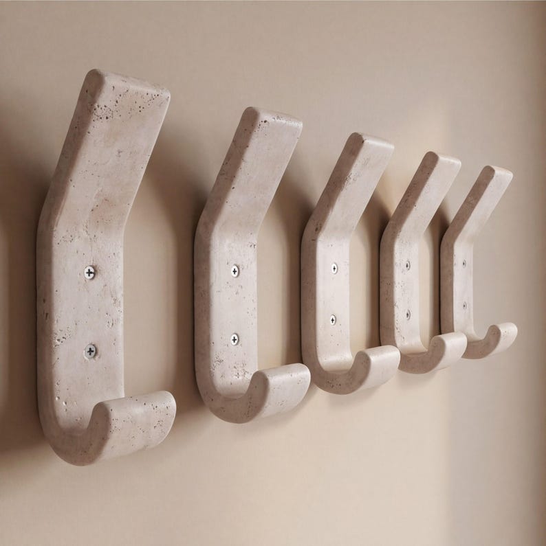 May include: Five off-white concrete wall hooks mounted on a beige wall. Each hook has a curved design with a rounded hook at the bottom and is secured with two screws. The hooks are evenly spaced.
