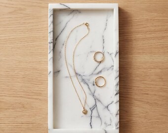 May include: A rectangular marble tray with a gold necklace and two gold rings. The necklace has a small pendant. The tray has gray veining and sits on a wooden surface. Jewelry.