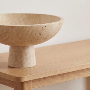 Puede incluir: Un cuenco de piedra beige con base de pedestal. El cuenco tiene un dise&ntilde;o ancho y poco profundo y se asienta sobre una base cil&iacute;ndrica corta. La superficie del cuenco y la base tiene un aspecto texturizado y poroso. El cuenco est&aacute; colocado sobre una superficie de madera clara.