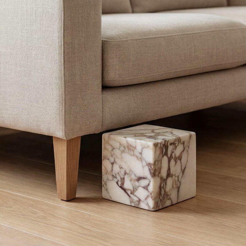 May include: A beige fabric sofa with wooden legs. A square marble side table with brown veining sits on the wooden floor next to the sofa. The table is a cube shape.