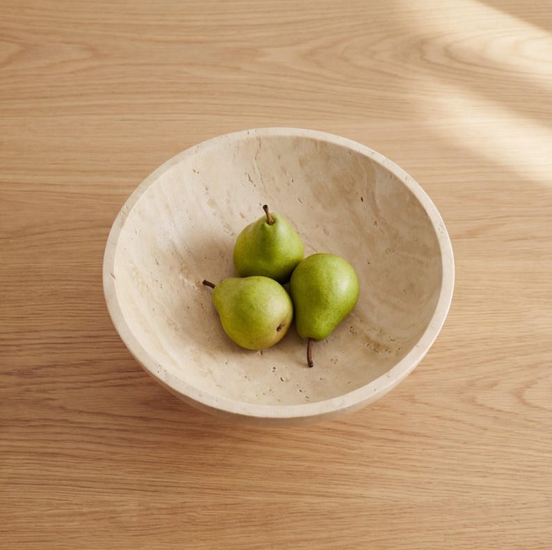Puede incluir: Un cuenco redondo de piedra beige contiene tres peras verdes. El cuenco tiene una superficie texturizada y descansa sobre una mesa de madera marr&oacute;n claro. Las peras tienen tallos marrones y est&aacute;n maduras.