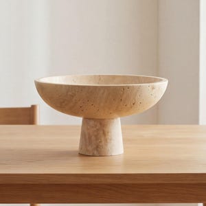 Puede incluir: Un cuenco de pedestal de piedra beige con una base ancha y poco profunda. El cuenco se asienta sobre una base cil&iacute;ndrica corta. La superficie tiene un aspecto texturizado y poroso. El cuenco se exhibe sobre una mesa de madera de color claro.