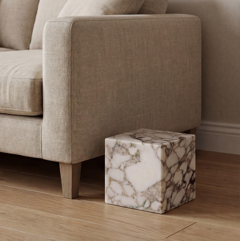 May include: A cube-shaped marble side table with a white and brown veined pattern. The table is next to a beige sofa. The table sits on a wooden floor. The table's dimensions are approximately 40cm x 40cm x 40cm.