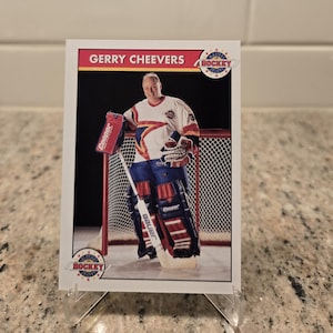 Peut inclure: Une carte de hockey représentant Gerry Cheevers en tenue de gardien de but. La carte montre Cheevers dans un maillot blanc, bleu et rouge, tenant une crosse de hockey. La carte porte le texte "Gerry Cheevers" et un logo d'équipe.
