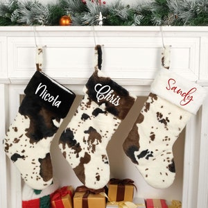 May include: Three personalized Christmas stockings with a cowhide pattern. The stockings are hung on a white fireplace mantel. The names "Nicola", "Chris", and "Sandy" are embroidered on the stockings.