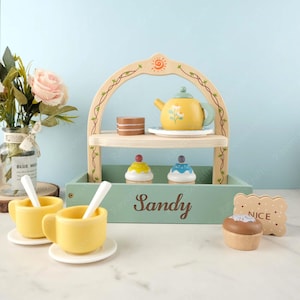 May include: A wooden toy tea set with a two-tiered stand, a teapot, two cupcakes, two teacups, two spoons, and a biscuit. The stand has a green base with the name "Sandy" written on it. The biscuit has the word "NICE" written on it.
