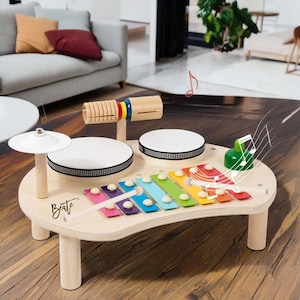 May include: A wooden musical toy set for children. The set includes a xylophone with colourful keys, two drums, a cymbal, and a wooden percussion instrument. The word "Beate" is written on the surface.